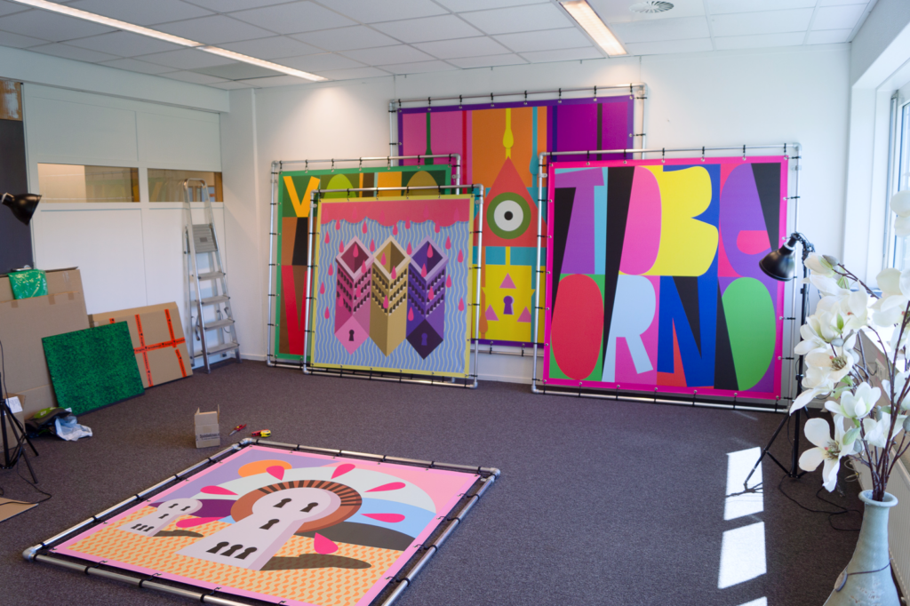 View of the studio of artist Ingmar Spit with large inkjet prints stacked together leaning on a wall. We see "Dead of the Dead" on the floor, "Toberlone", "Die Welt weint", "YOLO" and "Tagtigkeit der Hypophyse" leaning against the wall.