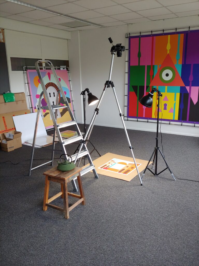 View of the studio of artist Ingmar Spit. We see a photographic setup taking pictures of work on the floor. We see a large inkjet print hanging on the wall called "Tatigkeit der hypophyse" and "Day of the dead"leaning on a wall.