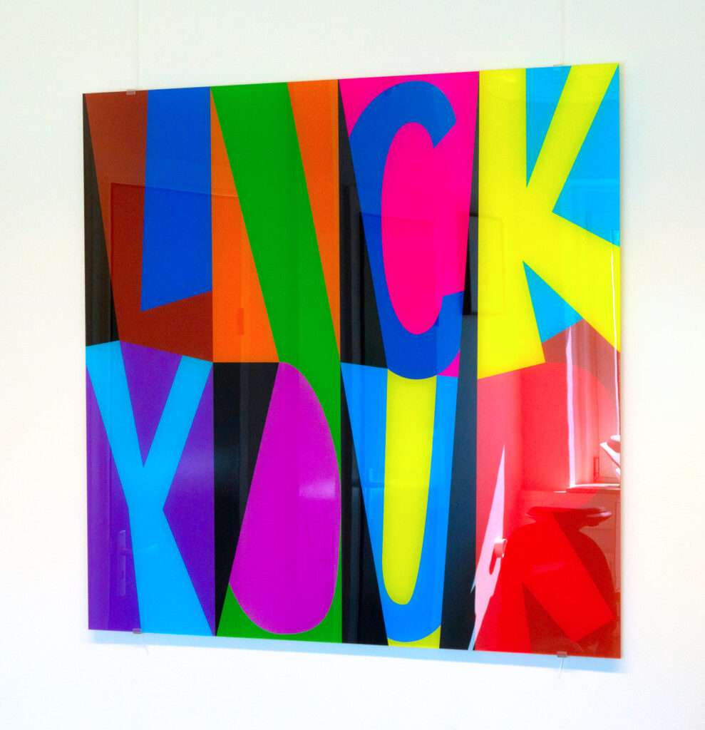 A square artwork titled 'Lick', printed on acrylic, is hanging on a white wall, suspended by two transparent acrylic cables. The word 'LICK' is crafted from bold, colourful, abstract shapes.