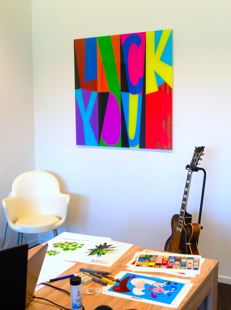 A square artwork titled Lick, inkjet-printed on acrylic, hangs on a white wall, suspended by two transparent cables. The words 'LICK' and 'YOUR' are made up of bold, colourful, abstract shapes.